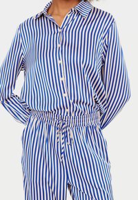 Person som bär en blå och vit vertikalt randig skjorta med knappar och matchande elastiska byxor med dragsko, med händerna vilande bakom.