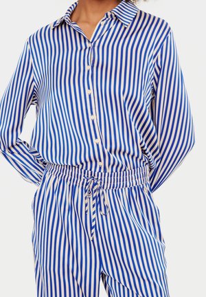 Persona vestida con una camisa de botones de rayas verticales azul y blanca y pantalones elásticos a juego con cintura ajustable, con las manos descansando en la espalda.