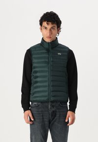 Zeleni pufast prsluk s visokim ovratnikom, zatvaranjem na zip i horizontalnim rebrastim dizajnom. Nosite preko crne majice s dugim rukavima. Vidljiv Lacoste logo.