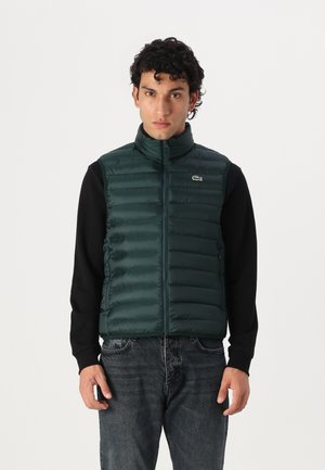 Gilet rembourré vert avec un col haut, fermeture éclair et design matelassé horizontal. Porté sur une chemise noire à manches longues. Logo Lacoste visible.