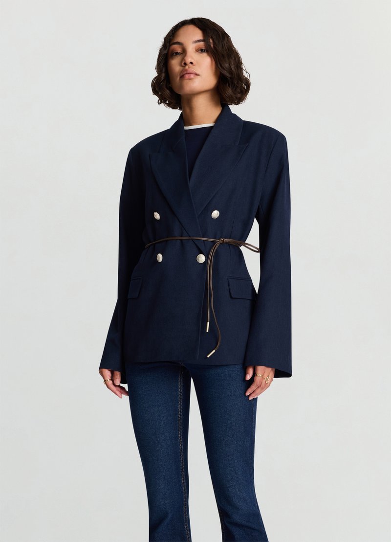 Blazer croisé bleu marine avec taille ceinturée, doté de boutons argentés, larges revers et poches à rabat. Tissu texturé, coupe structurée.