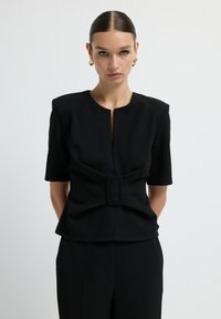 Top nero con maniche corte, scollatura rotonda e apertura centrale frontale. Presenta un design strutturato con un evidente dettaglio a fibbia.