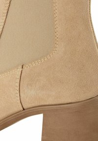 Beige semišové kotníkové boty s elastickými bočními panely, texturovaným povrchem a vrstveným dřevěným podpatkem. Obsahují viditelné švy podél švů.