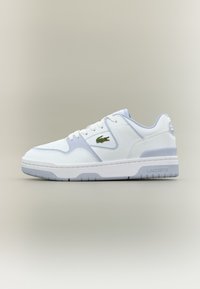 Λευκό και ανοιχτό γκρι αθλητικό παπούτσι Lacoste με πράσινο λογότυπο κροκοδείλου στο πλάι, λευκά κορδόνια και λαστιχένια σόλα σε ουδέτερο φόντο.