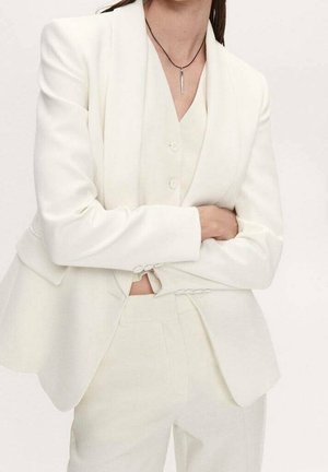 Mujer que lleva un traje blanco a medida con chaleco, blazer y pantalones, con los brazos cruzados y un collar con colgante de plata en un cordón negro.