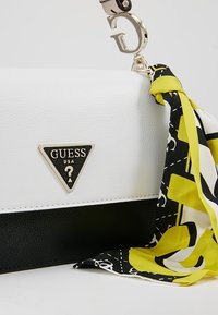 Borsa Guess bianca e nera con logo triangolare e una sciarpa a fantasia nera, bianca e gialla legata all'hardware dorato.