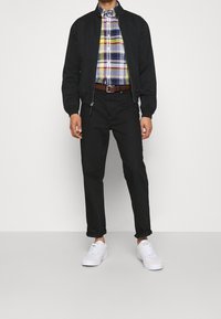 Svart bomberjacka över en flerfärgad rutig skjorta, svarta byxor och vita sneakers. Slät tyg, välsytt passform, dragkedjefickor, brunt bälte.