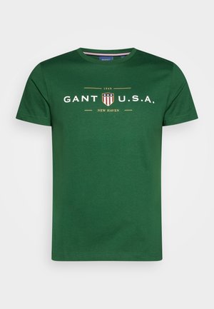 Grøn T-shirt med korte ærmer og rund hals med hvid tekst "GANT U.S.A." og et rødt, hvidt og blåt skjoldlogo med "1949" og "NEW HAVEN."