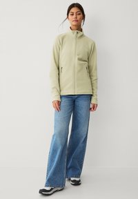 Giacca in pile con zip di colore verde chiaro, dotata di colletto alto, maniche lunghe e due tasche laterali con zip. Abbinate a jeans blu a gamba larga.