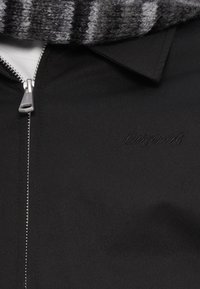 Μαύρο μπουφάν με φερμουάρ και υφή, με ασημί φερμουάρ και κεντημένο λογότυπο "Originals". Συνοδεύεται από γκρι ριγέ κασκόλ.