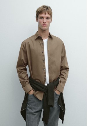 Chemise marron à boutons avec un t-shirt blanc en dessous, jean gris foncé, et un pull en maille foncé noué autour de la taille. Design simple, sans motifs.