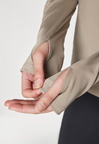 Haut ajusté à manches longues en tissu beige clair, avec des trous pour les pouces dans les manches, une texture lisse, une coupe élastique et des détails de couture contrastants.