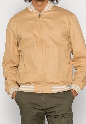 Homme portant une veste bomber beige zippée avec des poignets et un ourlet rayés, mains dans les poches, associée à un pantalon vert olive.