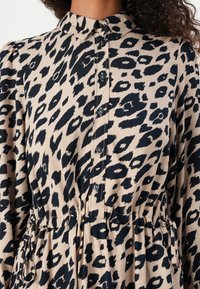 Kjole med leopardmønster som har en beige bakgrunn med svarte flekker, kragehals, knappelukking og justerbar snøring i livet.