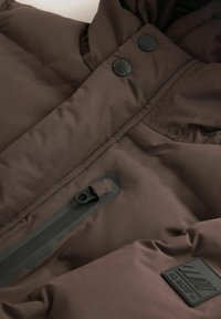 Doudoune marron avec une texture lisse, des boutons-pression noirs, une poche zippée et une étiquette en tissu.