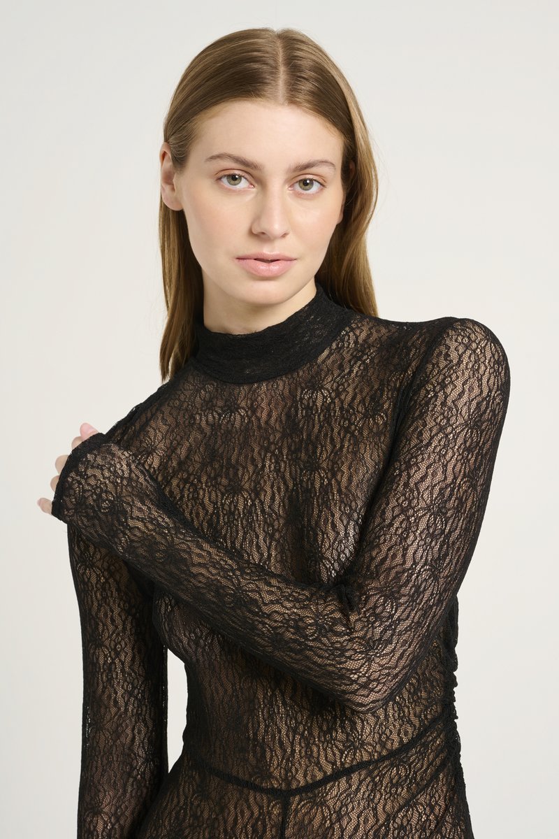 Top de encaje negro con mangas largas y cuello alto. Presenta intrincados patrones florales y una textura transparente. Ajuste ceñido al cuerpo.
