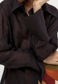 Czarna, półprzezroczysta koszula z długim rękawem, o klasycznym kołnierzyku i mankietach zapinanych na guziki. Miękka tekstura i luźny krój, przedstawiona na kolorowym tle.