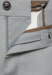 Pantalon en lin gris avec fermeture par bouton, poche zippée et surpiqûres marron en accent. Le tissu présente une texture apparente.