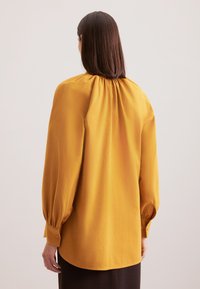 Senfgelbe Bluse mit langen Ärmeln und gerafftem Nähen am Hals sowie Knopfmanschetten. Weicher Stoff, lockere Passform, abgerundeter Saum.