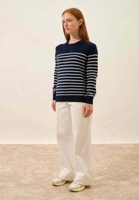 Granatowy i biały sweter w paski z okrągłym dekoltem, noszony z szerokimi białymi spodniami i beżowymi sneakersami, na jednolitym tle.