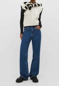 Gilet en maille torsadée épaisse couleur blanc crème porté sur un top à manches longues noir, associé à un jean pattes d'éléphant bleu et des bottes noires à semelle épaisse.