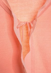 Indumento color pesco con superficie testurizzata, caratterizzato da un colletto ondulato e un dettaglio a fiocco al centro. Tessuto morbido, design casual.