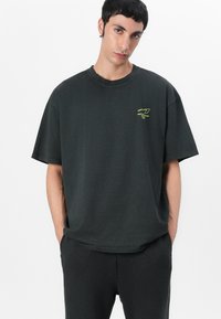Svart oversized bomull T-skjorte med rund hals og korte ermer, med en liten grønn logo på venstre brystområde.