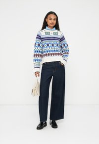 Maglione lavorato a maglia con motivi geometrici blu, verdi e bordeaux, abbinato a jeans larghi scuri e scarpe nere, mentre tiene una borsa color crema.