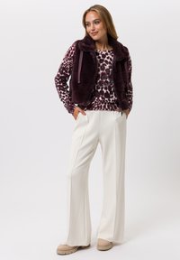 Nepbontvest in dieppaars over een top met patroon in tinten paars en crème. Witte wijde broek en beige platform schoenen maken de look compleet.