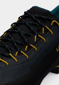 Μαύρο και γκρι παπούτσι αναρρίχησης με υφή στο πάνω μέρος, κίτρινες και τυρκουάζ κορδόνια, και ενισχυμένη σόλα για κράτημα και υποστήριξη.