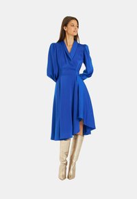 Blaues Wickelkleid mit Puffärmel, asymmetrischem Saum und glatter Textur. Kombiniert mit metallisch-silbernen kniehohen Stiefeln.