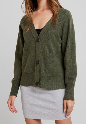 Kvinde iført en mørkegrøn cardigan med knapper og forlommer over en sort top og et lyst gråt skørt mod en ensartet baggrund.