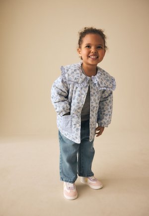 Smiling toddler with curly hair wearing a blue floral jacket with large collar, blue jeans, and pink sneakers against beige background.