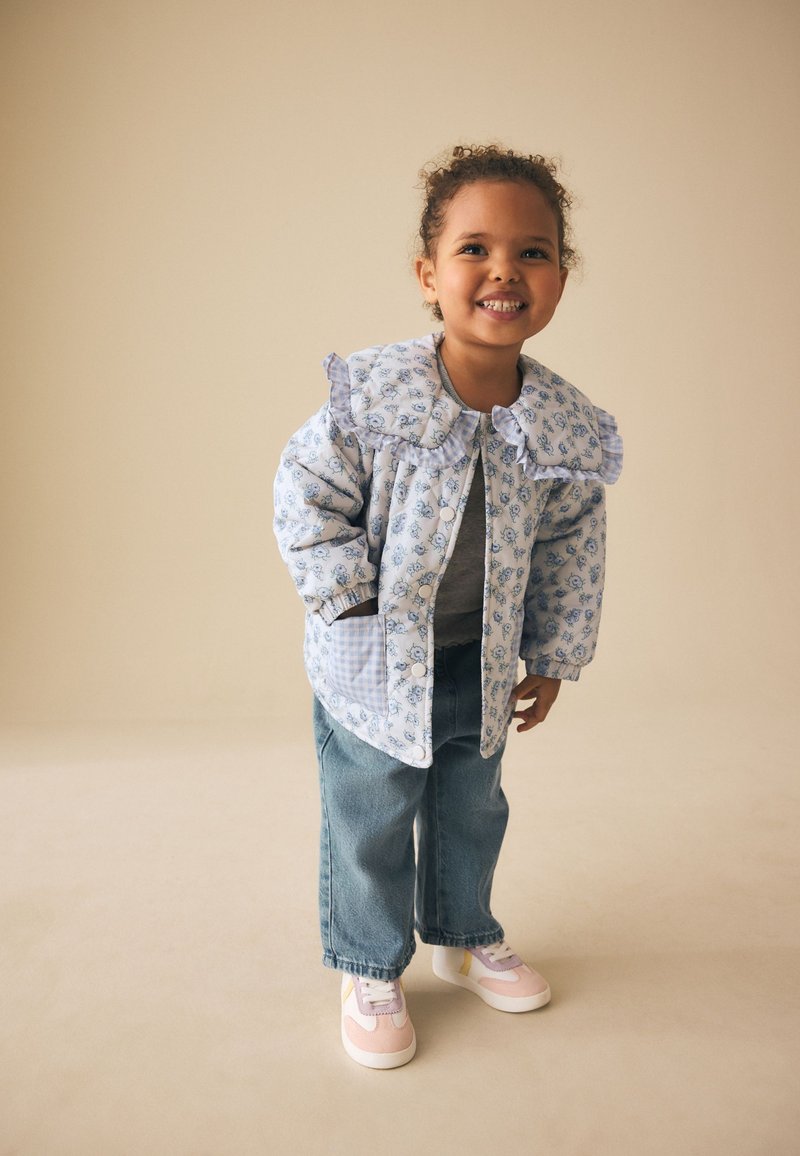Tout-petit souriant aux cheveux bouclés portant une veste bleue à fleurs avec un grand col, un jean bleu et des baskets roses sur fond beige.