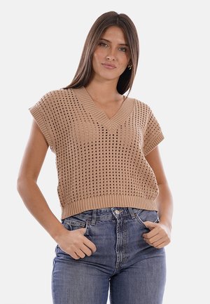 Donna con lunghi capelli castani che indossa un maglione beige lavorato a maglia senza maniche con scollo a V e jeans blu con le mani in tasca.
