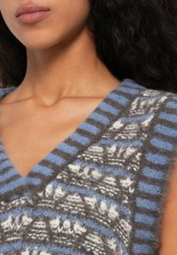 Primer plano de una persona que lleva un chaleco de punto esponjoso con patrones y rayas en azul, gris y blanco, mostrando el cuello y parte de la cara.