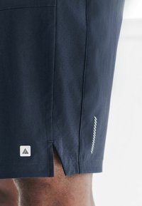 Granatowe spodenki sportowe, z perforowaną wstawką, bocznym rozcięciem i naszywką z logo w pobliżu dołu. Gładki materiał z czystym wzornictwem.