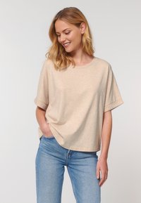 Persona sonriente con cabello rubio que lleva una camiseta beige clara y jeans azules, de pie frente a un fondo liso, con las manos en los bolsillos.