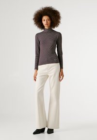 Naine, kellel on lokkis juuksed, kannab triibulist kõrge kaelusega pluusi, kreemja värvi laiad püksid ja musti kontsakingi, taustal hele ühtlane taust.