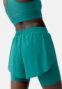 Teal sports bra with a fitted design, paired with loose teal shorts featuring a layered cut and elastic waistband. Smooth fabric texture.