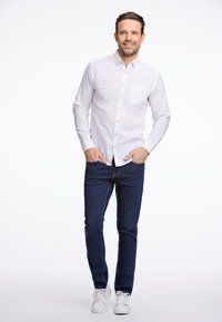 Camicia bianca a maniche lunghe con chiusura a bottoni e taschino sul petto, abbinata a jeans blu scuro e sneaker bianche, in piedi su uno sfondo chiaro.