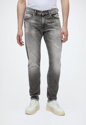 Muškarac u izblijedjelim sivim slim-fit trapericama s blagim oštećenjima i bijelim tenisicama na jednobijeloj pozadini.
