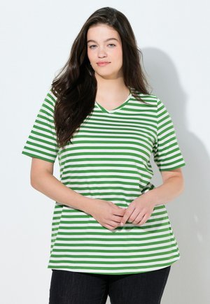 Giovane donna con capelli castani lunghi che indossa una maglietta a maniche corte verde e bianca a righe, in piedi davanti a uno sfondo chiaro uniforme.