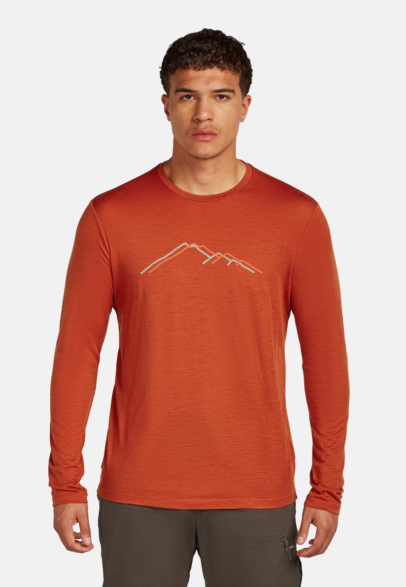 Junger Mann mit kurzen lockigen Haaren, der ein langärmeliges Hemd in rostfarbener Farbe mit einem minimalistischen Bergdesign auf der Brust trägt.