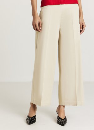 Beige weite Hosen aus glattem Stoff, mit klaren Falten und lockerem Schnitt, kombiniert mit spitzen schwarzen Schuhen, die Ösen-Akzente aufweisen.