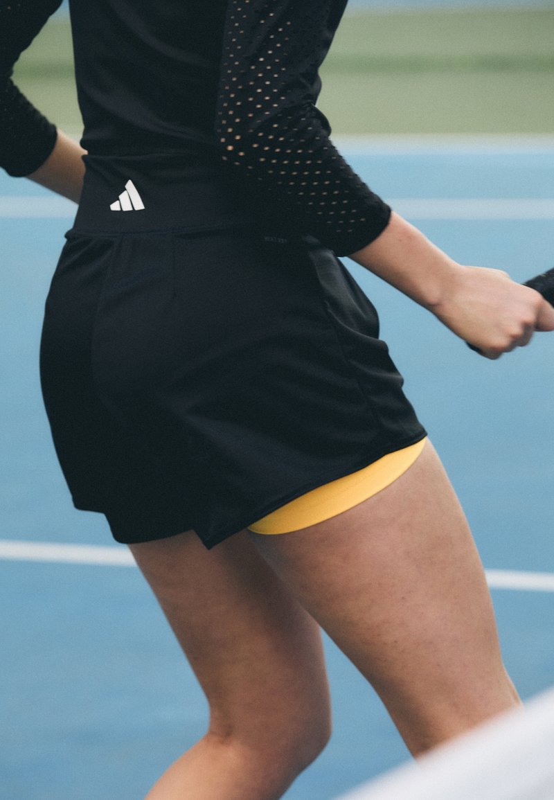 Svarta sportshorts med en gul innerbyxa. Funktioner inkluderar en stilren design och en logotyp på baksidan av midjebandet. Slät tygstruktur.