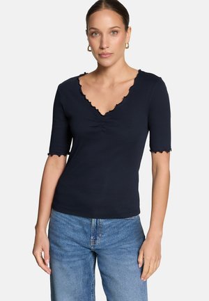 Femme portant un haut bleu marine ajusté avec un col en V festonné et des manches, associée à un jean bleu, debout devant un fond uni.