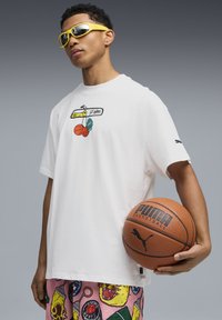 Witte t-shirt met een grafisch ontwerp dat een hanger, basketbal en een groene accent toont. Model houdt een bruine Puma basketbal vast en draagt gele zonnebril.