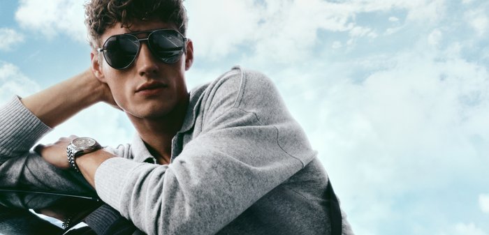 Ung mann med mørke solbriller og en grå genser, hviler armen på en reflekterende overflate med en delvis skyet himmel i bakgrunnen.
