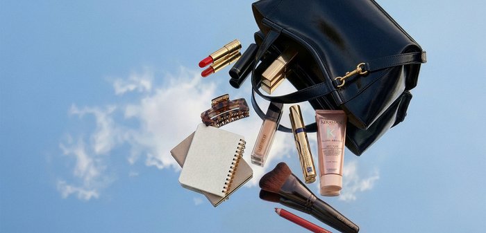 Sac à main noir avec rouge à lèvres, pinceau de maquillage, fond de teint, pince à cheveux, carnet et tube de soin capillaire sur fond de ciel bleu.